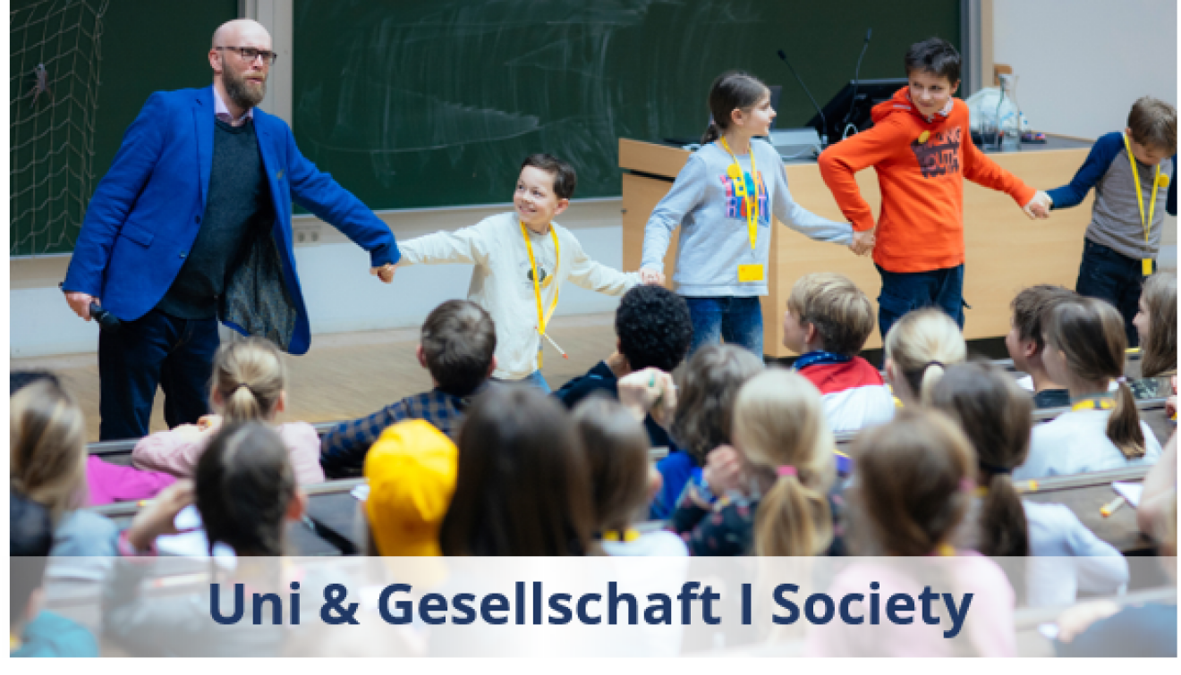 Vorlesung der Kinder-Uni: Dozent mit Bart und blauem Sakko in einer Reihe mit Kindern, davor Sitzreihen mit Kindern