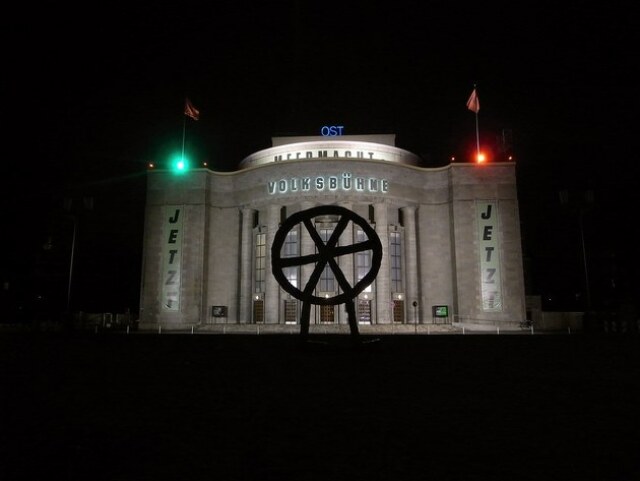 Foto der Volksbühne bei Nacht, im Vordergrund ist ein sechspeichiges Rad zu sehen. 
