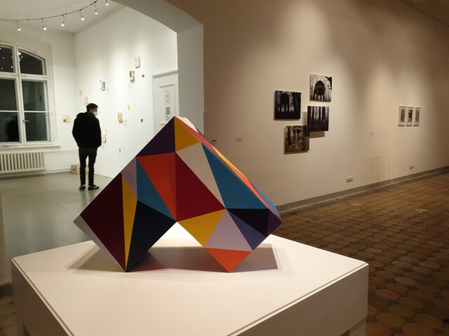 Bild der Ausstellung 'knoten' in der Galerie. Im Vordergrund ist eine bunte, geometrische Figur. Im Hintergrund ist ein Besucher sowie mehrere an der Wand hängende Bilder.