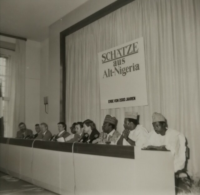 Das schwarz-weiß Foto zeigt einen langen Tisch, an dem 10 Personen sitzen. Dahinter ist eine Wand mit weißem Vorhang auf dem ein Schild hängt mit der Aufschrift 'Schätze aus Alt-Nigeria - Erbe von 2000 Jahren'.
