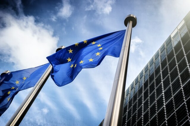 Im Hintergrund ist ein blauer Himmel mit weißen Wolken zu sehen. Rechts ist die Fassade einen Gebäudes angeschnitten dargestellt. Im Vordergrund stehen zwei Fahnenmasten an denen die EU-Fahne weht.