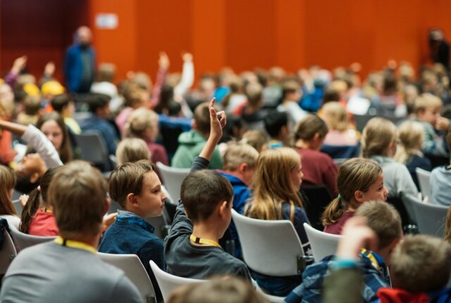 Foto eines Hörsaals in dem Kinder sitzen, einige Kinder heben die Hand.