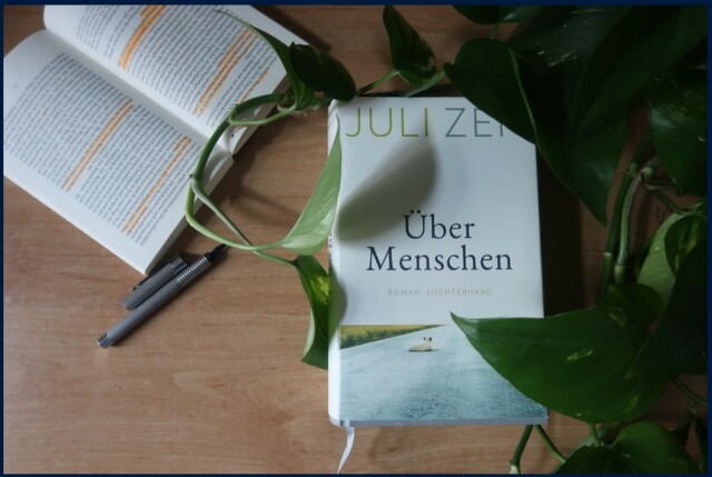 Foto des auf einem Tisch liegenden Romans 'Über Menschen' mit einer umringenden Pflanze.