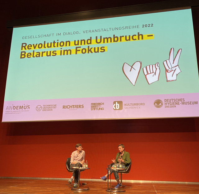 Foto zeigt die Aktivistin Olga Spharaga mit Anika Walke. Beide sitzen auf Stühlen auf einer Bühne vor einer roten Wand. Auf einer Leinwand über den beiden steht der Titel der Veranstaltung „Revolution und Umbruch – Belarus im Fokus“