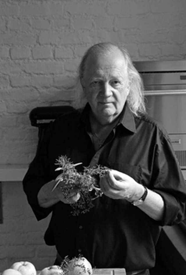 Foto von Jürgen Dollase, der in einer Küche steht, in die Kamera blickt und dabei Kräuter in der Hand hält.