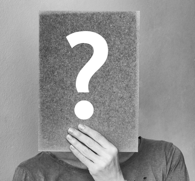 Ein schwarz-weißes Bild von einem Menschen, der eine Mappe mit Fragezeichen vor das Gesicht hält.