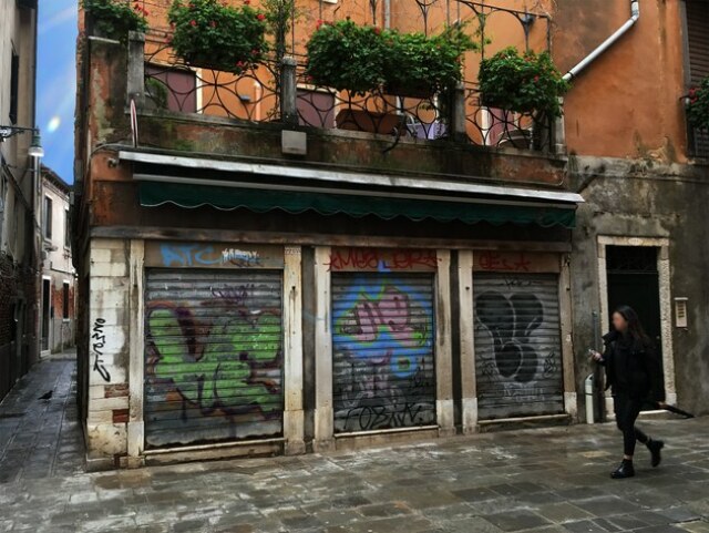 Foto eines Wohngebäudes. Im Zentrum sind heruntergezogene Rolltore, auf denen Graffitis gesprüht sind. Rechts läuft eine Frau durchs Bild.