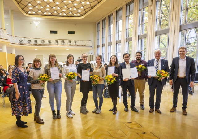 Foto Preisverleihung. 11 Personen mit Urkunden und Blumen im beleuchteten Saal.