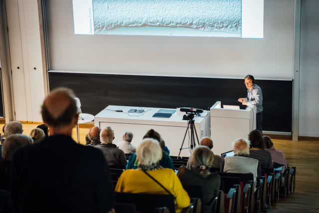 Prof. Dr. Marius Ader steht an einem Pult und redet vor einem Publikum in einem Hörsaal. Hinter ihm wird eine Präsentation per Beamer an die Wand projeziert. In der Mitte des Raumes steht eine Kamera zur Live-Übertragung des Vortrages.