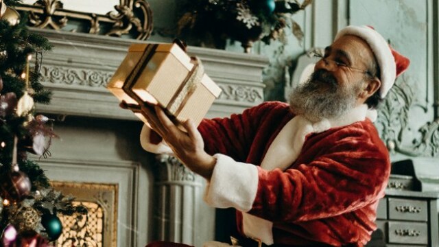 Der Weihnachtsmann hält ein Geschenk in der Hand. Er steht vor einem Kamin, am linken Rand ist ein Weihnachtsbaum zu sehen.