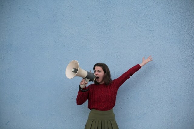 Eine Frau steht vor einer blauen Wand und schreit in ein Megaphon