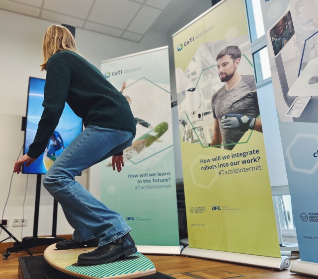 Picture of a girl trying out CeTI's Surfboard Demonstrator. She is standing on a spring-loaded surfboard in front of a screen and her movements are tracked by a camera and then compared with the virtual figure. 
