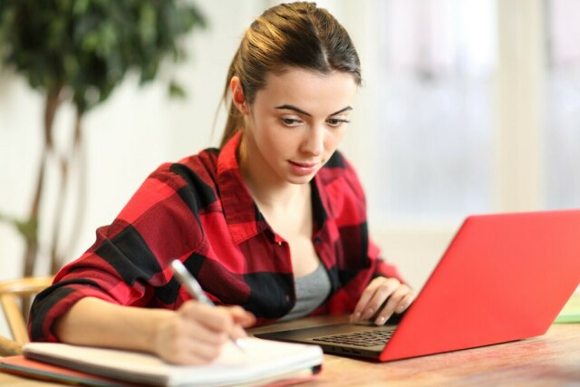eine Frau in rot-schwarz karierter Bluse sitzt am Tisch vor einen roten Notebook und schreibt Notizen auf einem Block