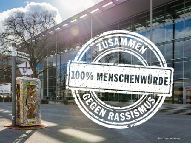 Foto vom HSZ der TUD von der Seite des Haupteinganges. Das Logo der Internationalen Wochen ist groß und zentral angelegt, auf dem 'Zusammen gegen Rassismus. 100 % Menschenwürde' steht.