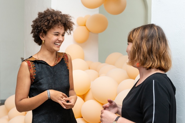 Foto, zwei junge Frauen lachend im Gespräch. Im Hintergrund helle Luftballons.