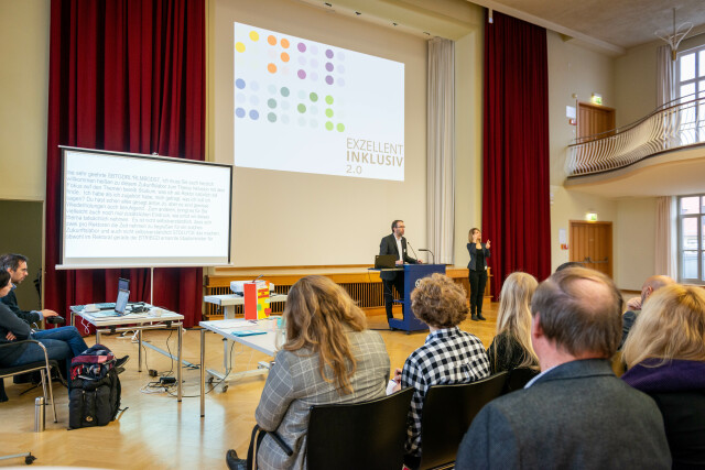 Foto zeigt einen Vortrag aus der Zuhörerperspektive im Dülfersaal der TU Dresden. Im Hintergrund erkennt man den Referenten und die Gebärdendolmetscherin vor einer Präsentation mit der Aufschrift: 'Exzellent Inklusiv 2.0'.