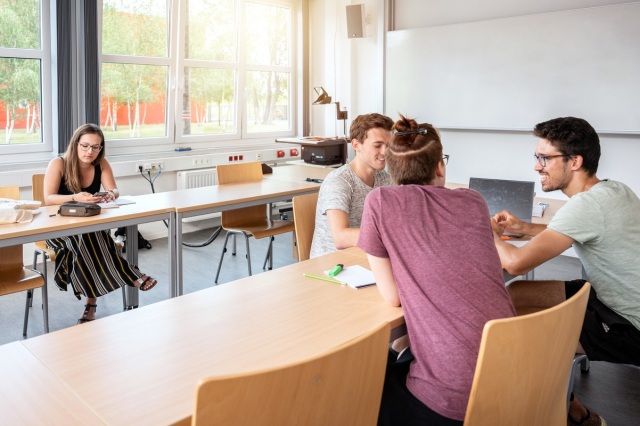 Vier Studierende sitzen in einem Seminarraum. Drei davon unterhalten sich, während eine Studentin an ihrem Handy sitzt.