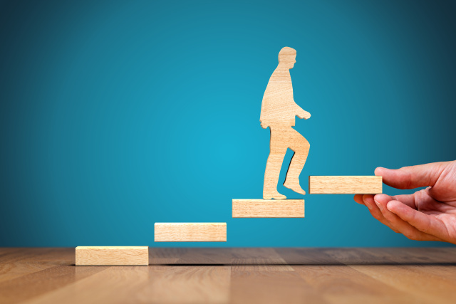 stylized figure climbs wooden stairs in front of a blue background; the last step is held by a human hand