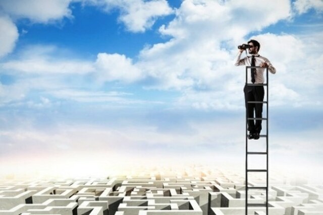männliche Person auf einer Leiter schaut mit einem Fernglas in die Ferne, im Hintergrund blauer Himmel und ein großes Labyrinth