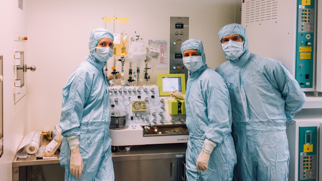 Drei Personen mit Schutzanzügen, Handschuhen, Masken und Hüten posieren vor einer Maschine in einer Laborumgebung. An der Maschine sind Infusionsbeutel angeschlossen.