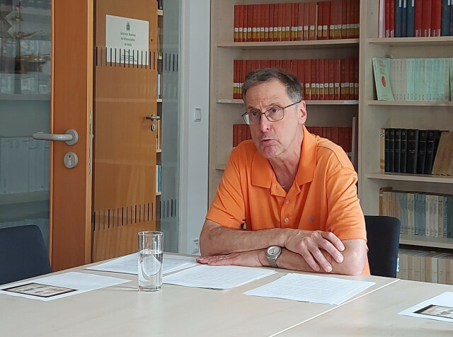The picture shows Bruce Brasington. He is wearing an orange polo shirt and sitting at a table with his arms crossed. He looks past the camera and speaks. His notes are lying on the table in front of him. In front of these notes is a glass of water.