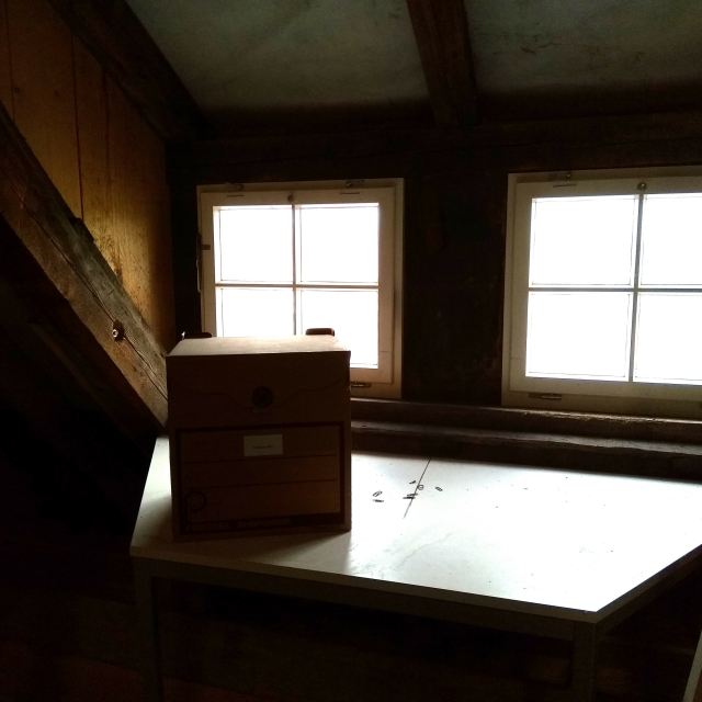 Fotografie eines Raumes mit Dachschrägen. Zu sehen ist eine dunkle Ecke mit zwei Fenstern, vor denen ein Tisch und ein Archivkarton platziert sind.