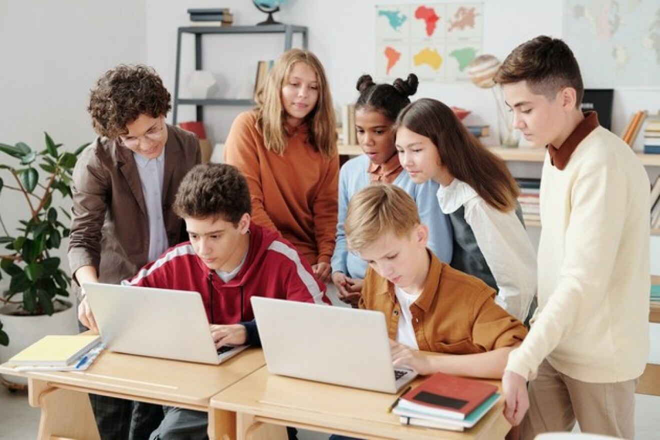 Eine Gruppe Schüler:innen steht/sitzt in einem hellen, freundlichen Klassenzimmer um zwei Laptops herum.