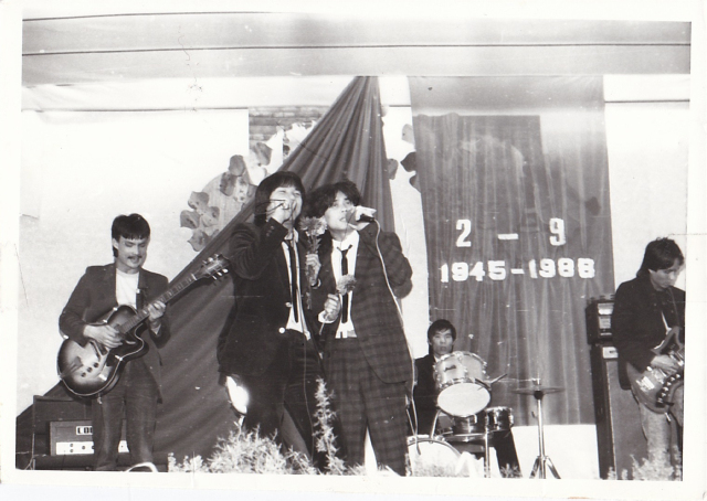 In the photo there are four musicians (2 singers, bass, guitar, drums). In the background hangs a large cloth on the wall, which says '2 - 9 1945-1988'.
