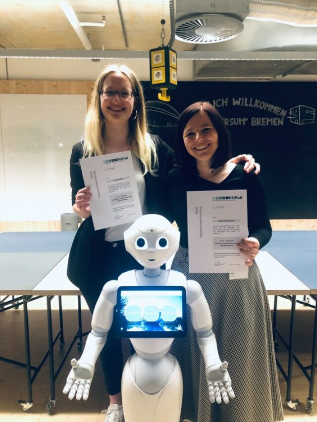 zwei Frauen mit Urkunden in der Hand lächeln in die Kamera, vor ihnen steht ein weißer Roboter