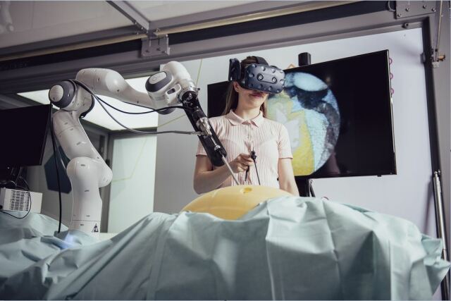 Eine Frau mit VR-Brille übt mit Hilfe eines Roboterarms eine Operation an einem Simulator. 