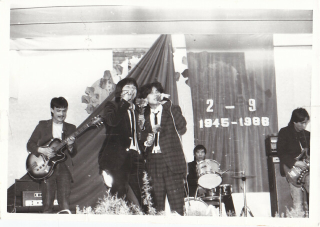 schwarz-weiß-Foto einer Band, auf der Bühne stehen zwei Personen mit Mikrophon in der Hand. Im Hintergrund steht ein Gitarrist und ein Schlagzeuger
