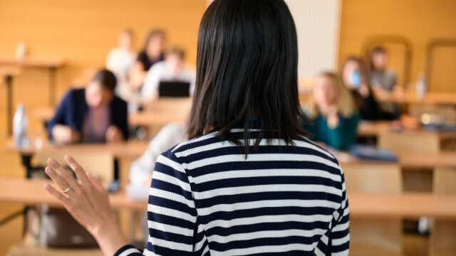 The photo shows a woman with shoulder-length dark hair from behind. She is wearing a black and white striped sweater and has her left hand raised to chest level. In the background you can see a seminar room with female listeners. 