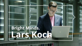 Prof. Koch ist im rechten Teil des Fotos. Er steht an einem Geländer, auf dem ein aufgeklappter Laptop steht, in den er hineinblickt. Im Hintergrund geschlossene Außenjalousien.