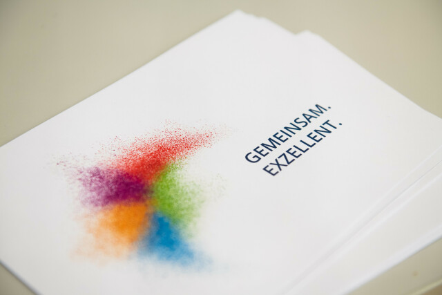 On a white card in A5 landscape format are different colored dots that form a swarm. To the right of the swarm are the words 'together excellent'.