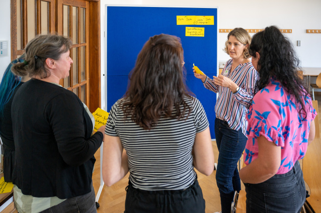 5 women stand around a metaplan board and discuss, holding yellow cards in their hands.