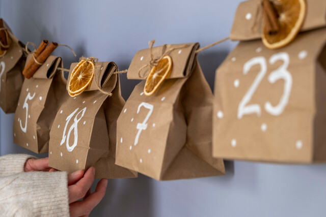 The photo shows part of an Advent calendar made from paper bags. The numbers 5, 18, 7 and 23 are written on the bags, which have dried orange slices attached to them.