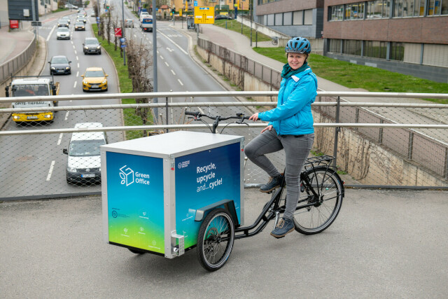 The photo shows a person sitting on a cargo bike. The person and the bike are standing on a bridge, with a road with cars below. 