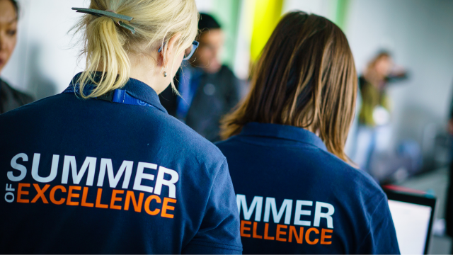 two female persons in back view, wearing blue T-shirts with the inscription: Summer of Excellence