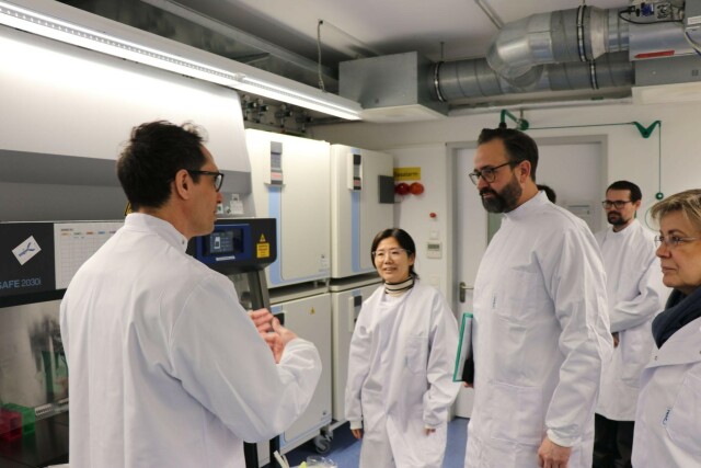 People standing in the laboratory wearing white labcoats. A man is in the front explaning to the rest of people