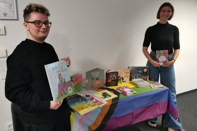 Zwei Personen stehen an einem Tisch gefüllt mit Schulbüchern für queere Grundschulkinder