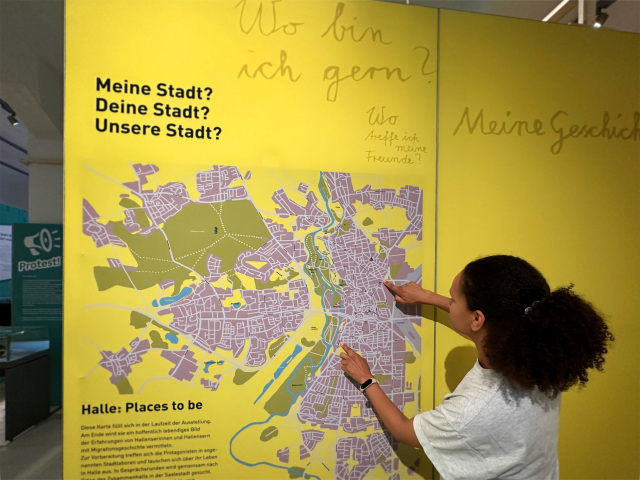 Eine Person vor einer gelben Wand mit einer Stadtkarte von Halle. Text auf der Wand: 'Meine Stadt? Deine Stdt? Unsere Stadt?' u.a.