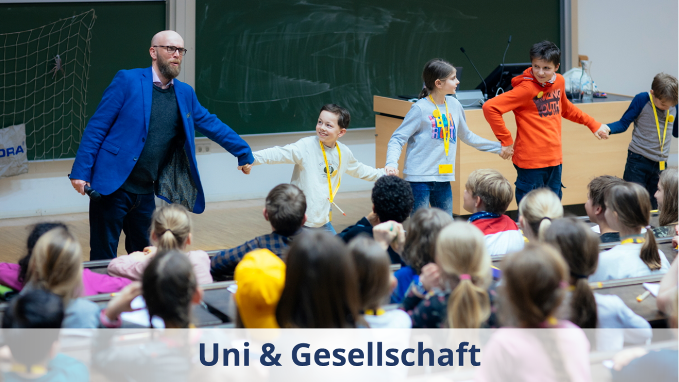 Das Foto zeigt eine Vorlesung der Kinder-Uni: Es zeigt einen Dozenten mit Bart und blauem Sakko in einer Reihe mit Kindern, davor sind Sitzreihen mit Kindern, die nach vorn schauen.