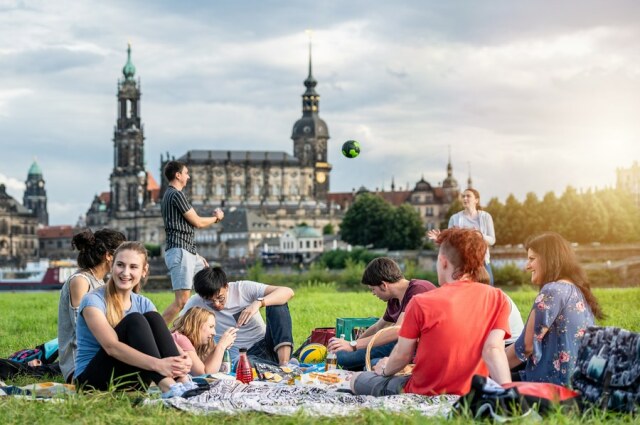 Young people sit together with a smile on a meadow against the backdrop of Dresden.
