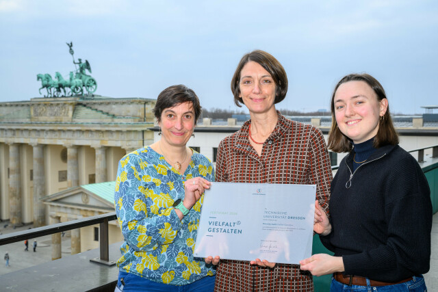 Photo of three women, in the middle Prorector University Culture holds the award in her hands. In the background the Brandenburg Gate, Berlin.