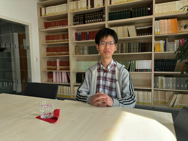 Hikaru Itoh sits at a table. In the background, one can see several book shelves.