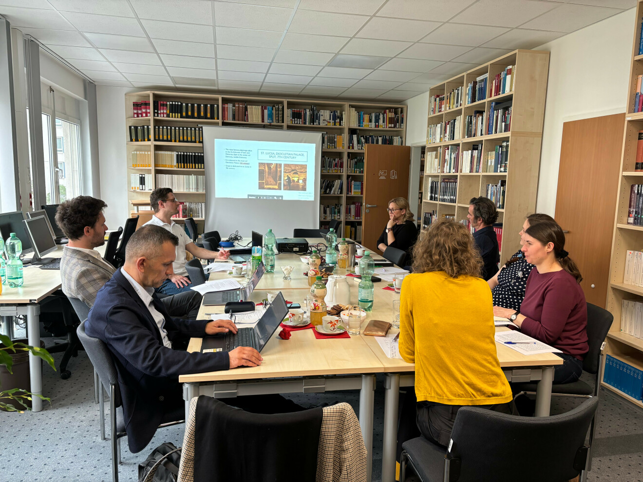 View of FOVOG library. Several people are sitting around a long table. In the background, a presentation is shown.