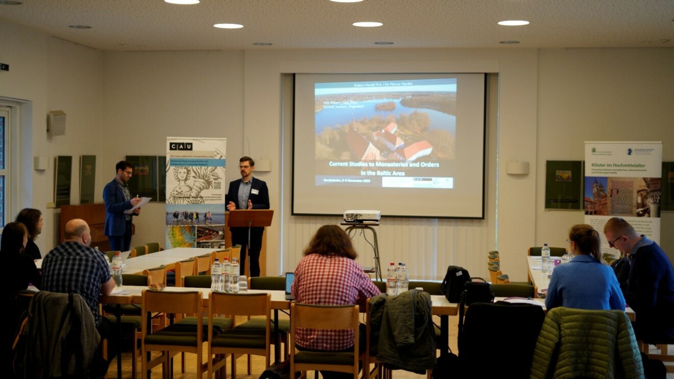 The photograph shows an event room from the rear perspective of the room. A screen can be seen in the center showing a presentation and two men, one of them is giving a lecture on the presentation. Furthermore, six people can be seen from behind, sitting 