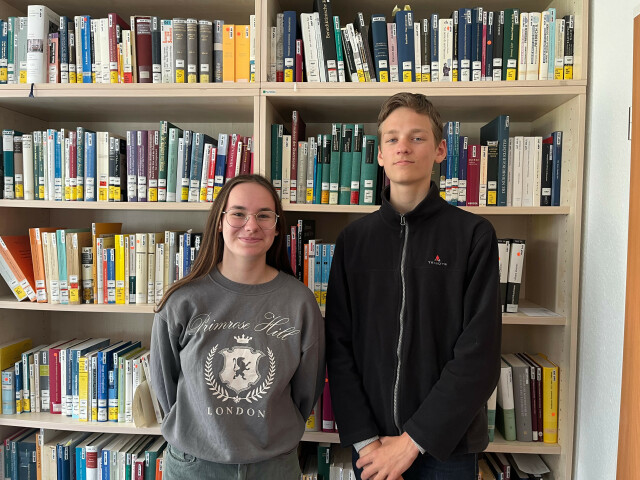 Two pupils, a boy and a girl, stand in front of a book shelf.