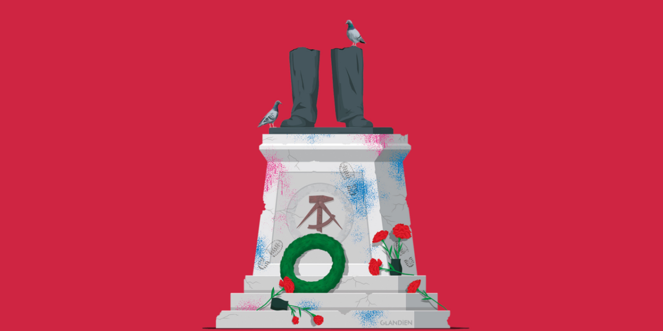 Vandalized pedestal of a monument with hammer and sickle, only the legs are left of the figure on the pedestal. Roses and a wreath lie untidily in front of it. 