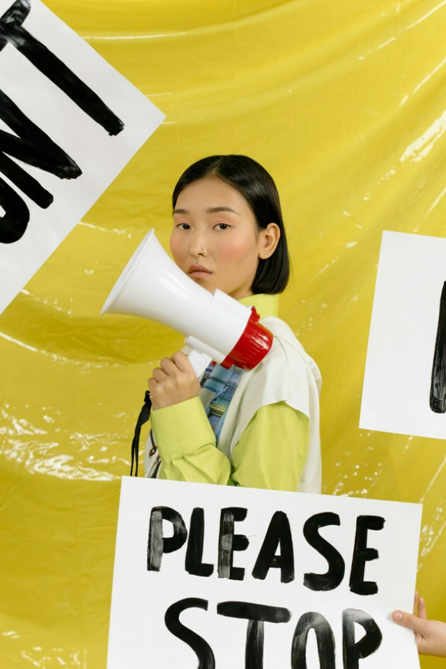 A woman holds a megaphone in her hand. She can be seen in profile. A sign reading 'Please stop' is held in front of her.